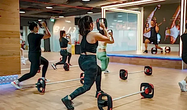 Members using Les Mills SMARTBAR™ equipment during a BODYPUMP class at GymNation, following the instructor on stage inside a fitness studio.